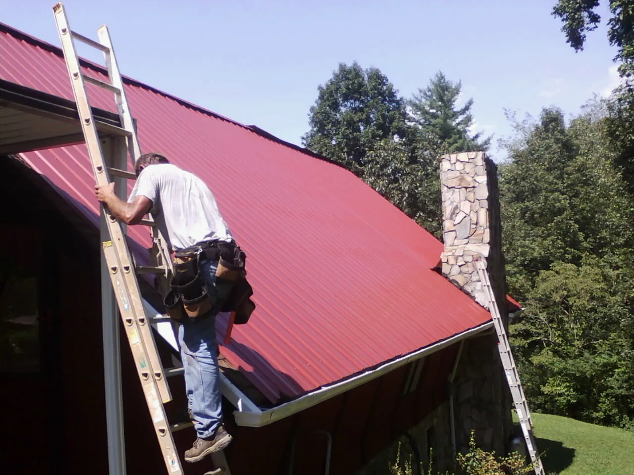 Roofer on a ladder beside a red metal roof and stone chimney — Soffit & Fascia Repair by Roofing Brothers in Framingham
